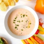 creamy paprika dip in a white bowl garnished with herbs and olive oil surrounded by crudites and chips.