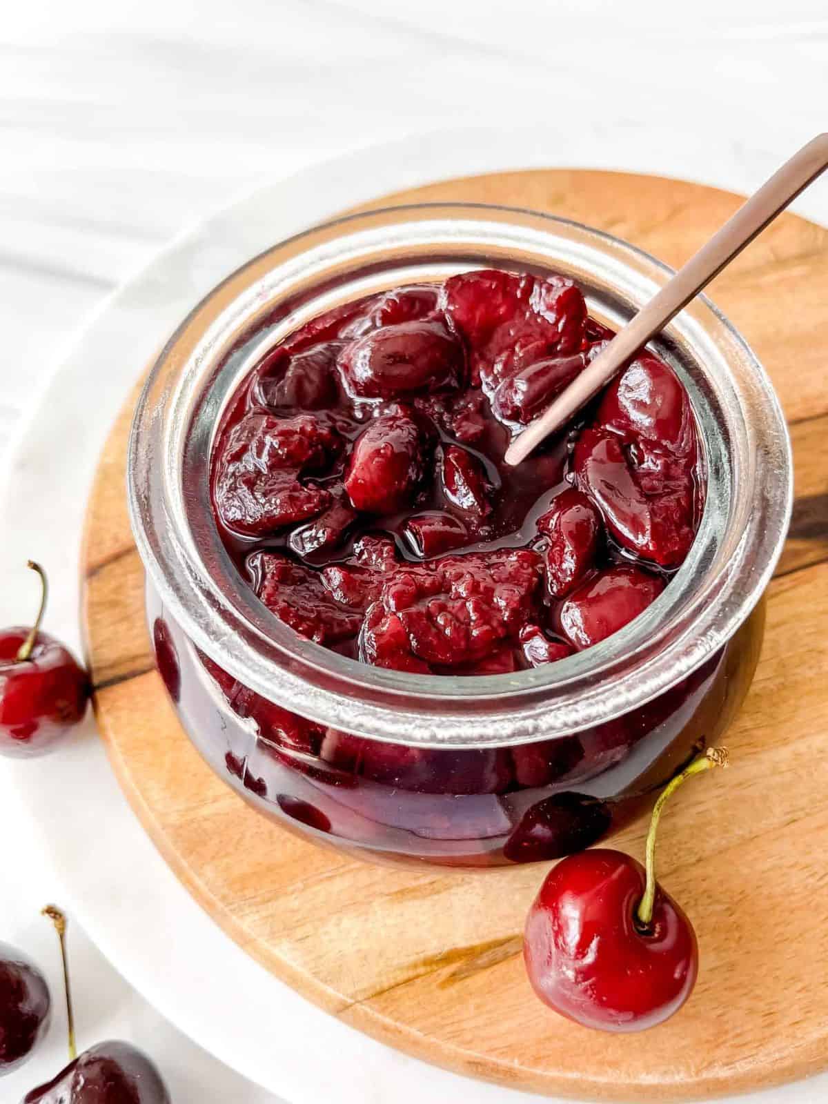 cherry compote in a glass jar with a spoon in it on a wooden board next to cherries.