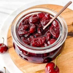 cherry compote in a glass jar with a spoon in it on a wooden board next to cherries.