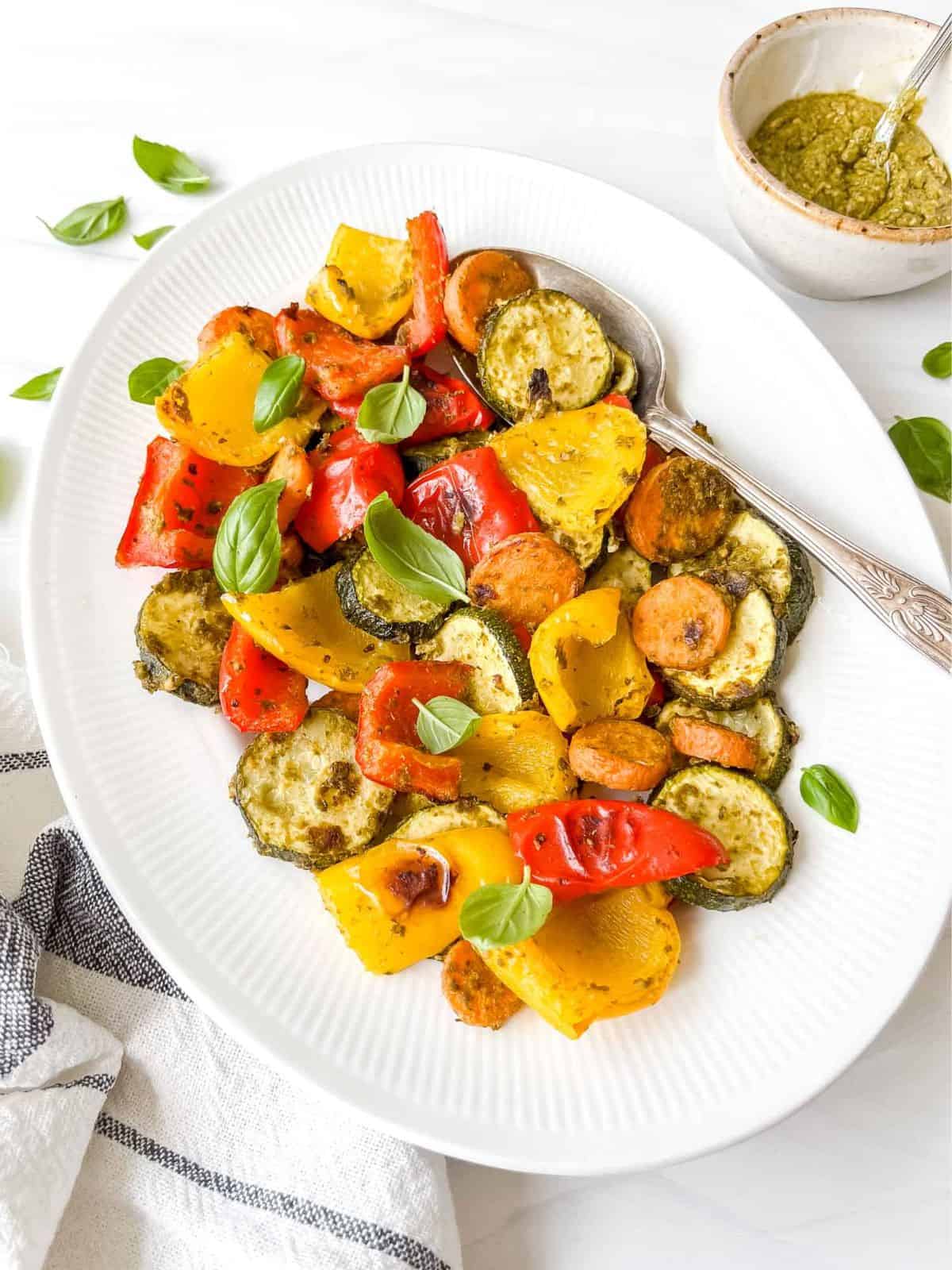 pesto roasted vegetables on a white oval plate next to a bowl of pesto with a spoon in it.