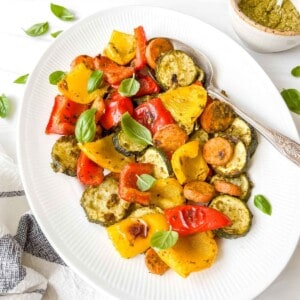pesto roasted vegetables on a white plate with a spoon on it next to a bowl of pesto and basil leaves.