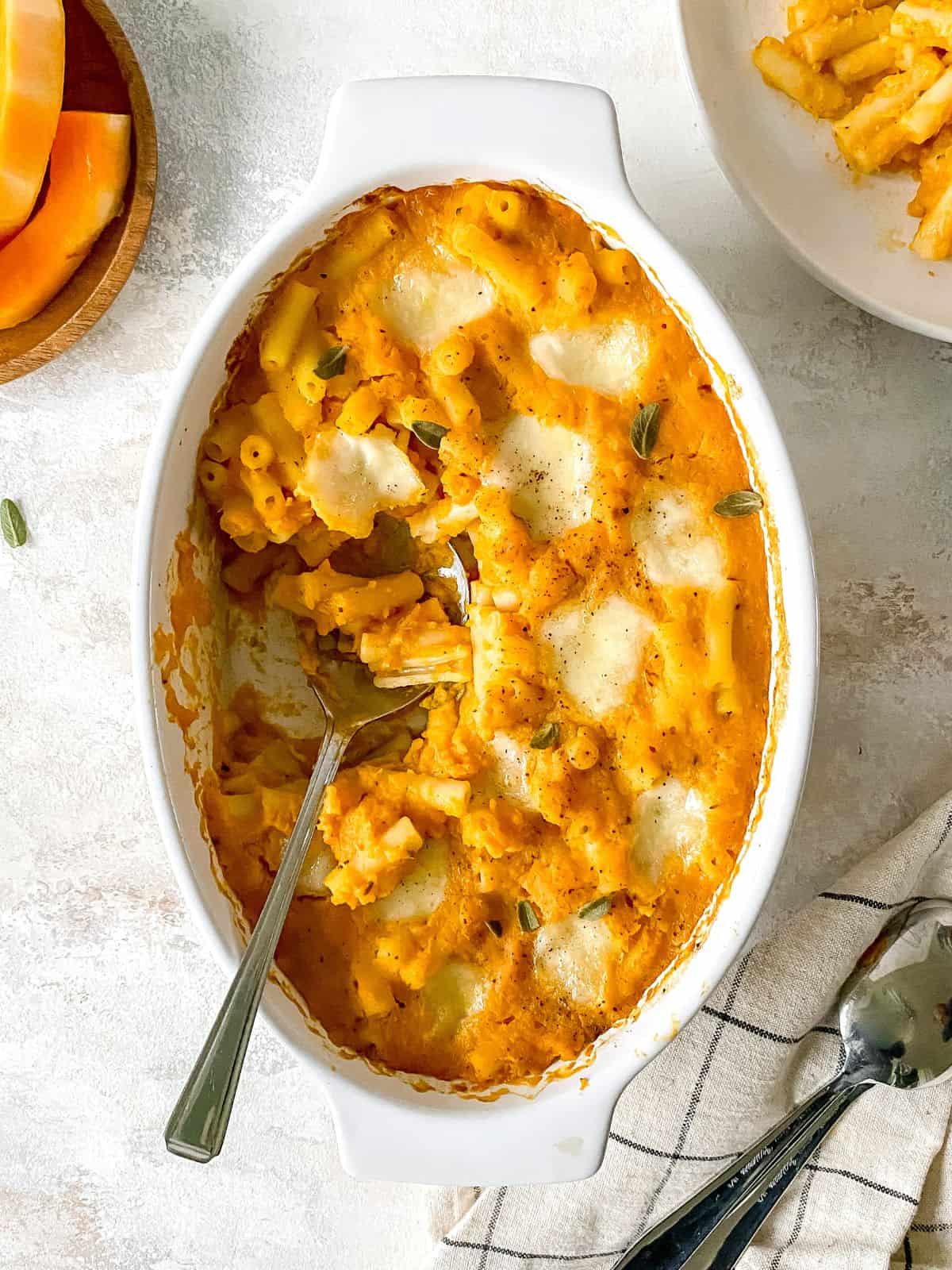 butternut squash pasta bake in a white dish with a spoon in it next to a plate of pasta and cutlery.