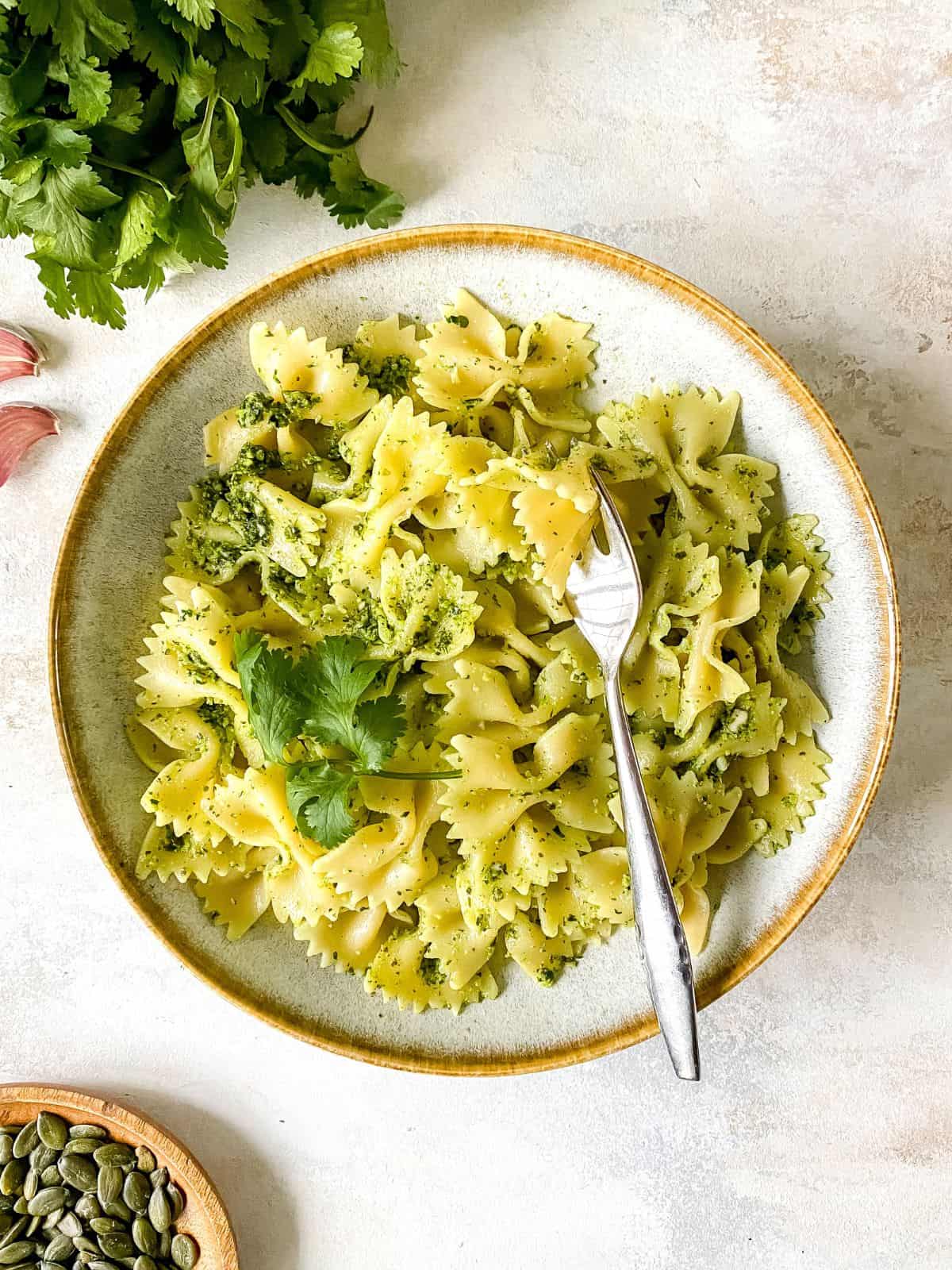coriander pesto pasta in a light grey bowl with a fork in it next to fresh herbs and pepitas.