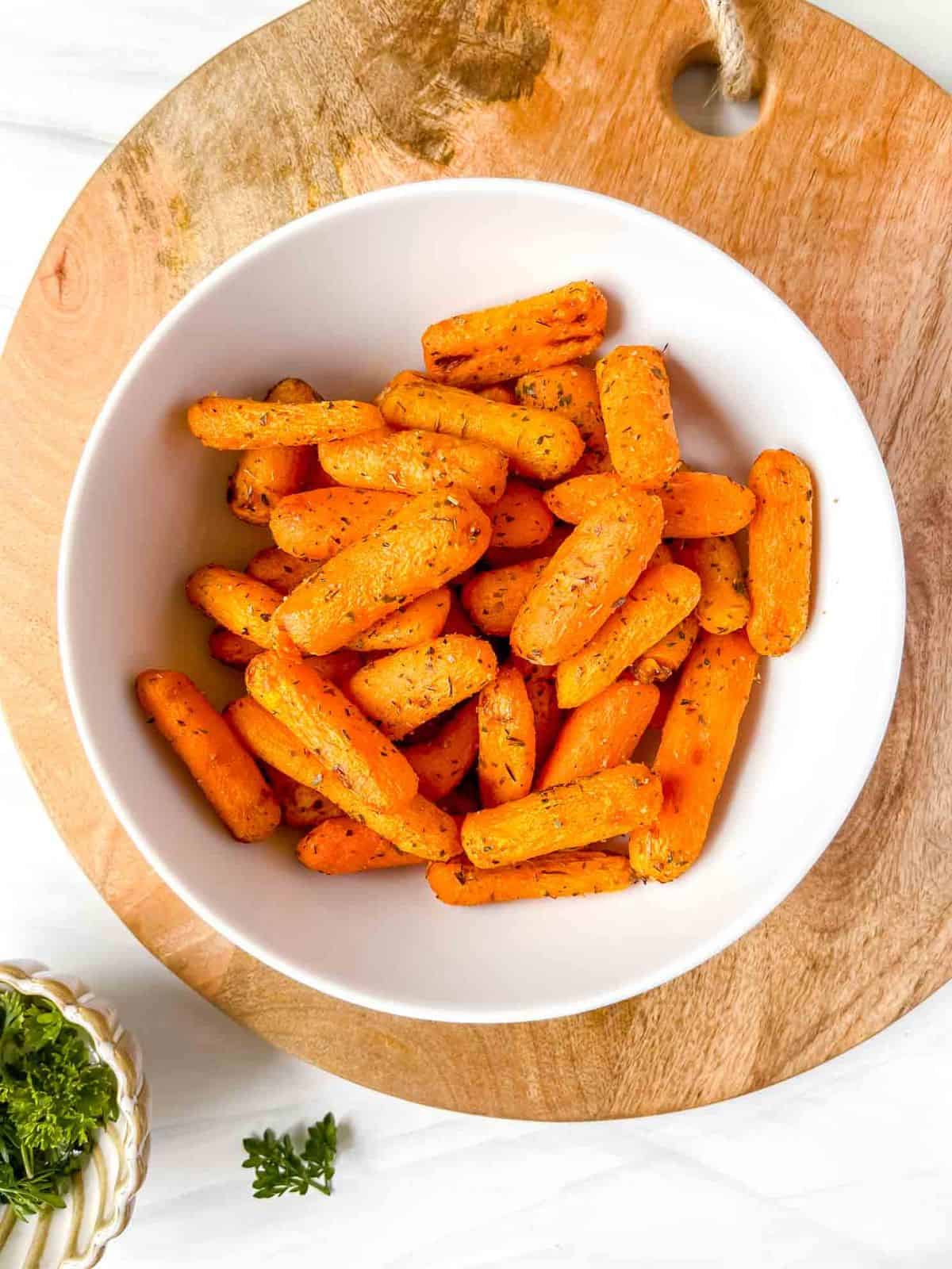 air fryer baby carrots in a white bowl on a wooden board next to a small bowl of herbs.
