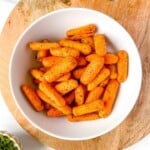 air fryer baby carrots in a white bowl on a wooden board next to a small bowl of herbs.