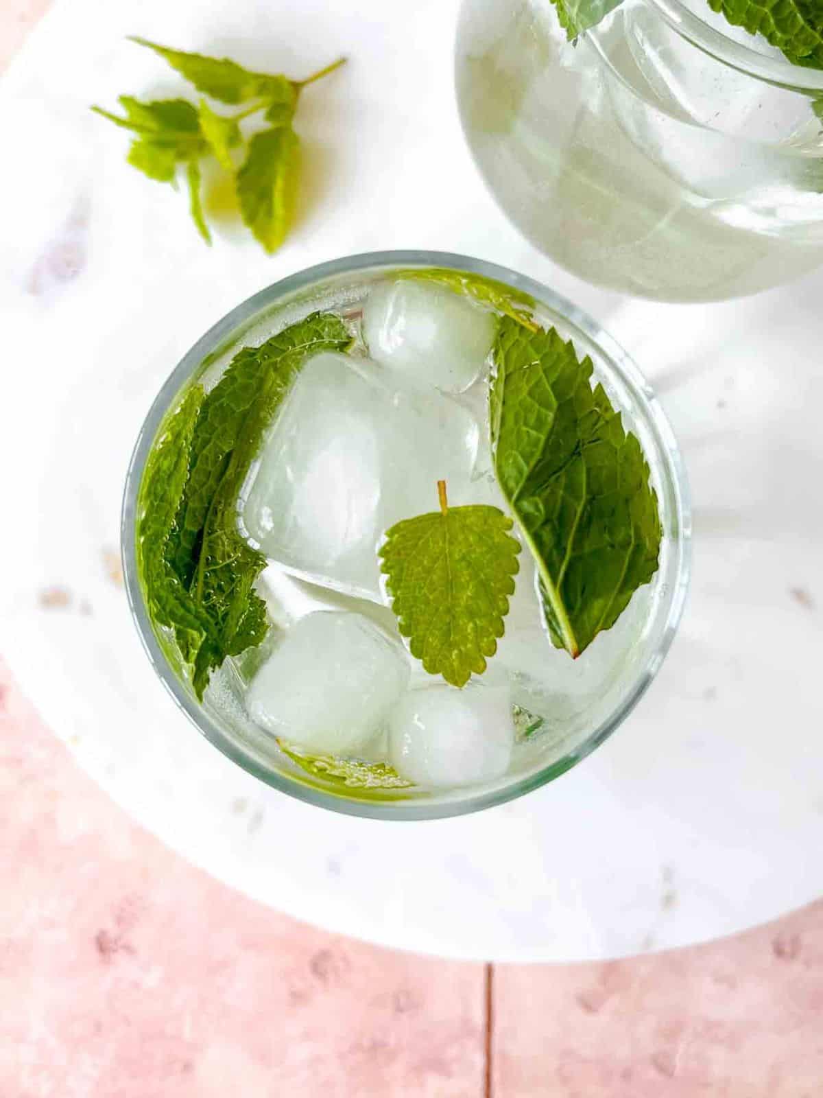 hugo spritz mocktail in two glasses on a marble board on a pink table next to lemon balm leaves.