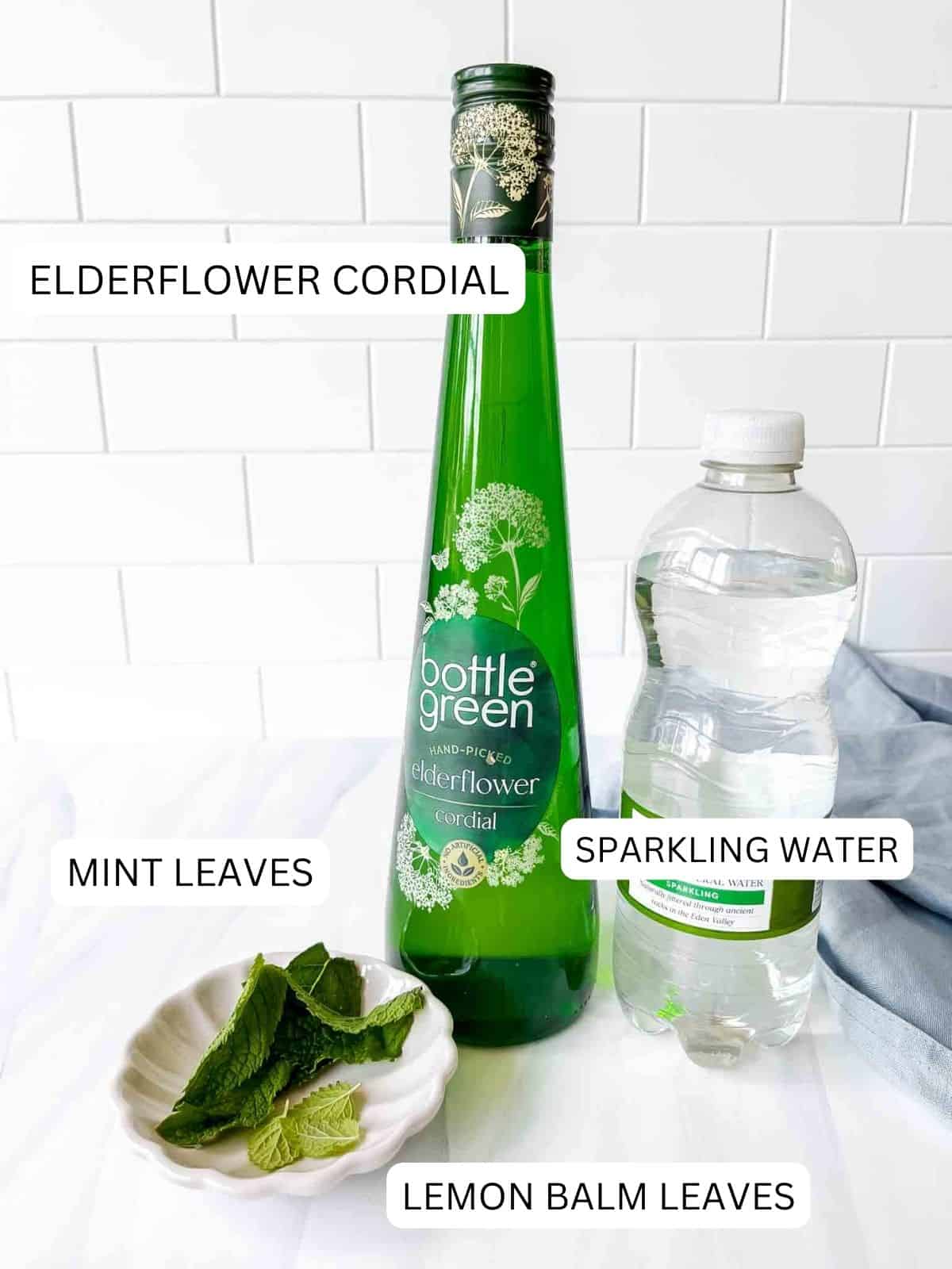 individually labelled bottles of elderflower cordial and sparkling water and a bowl of labelled mint leaves and lemon balm leaves.