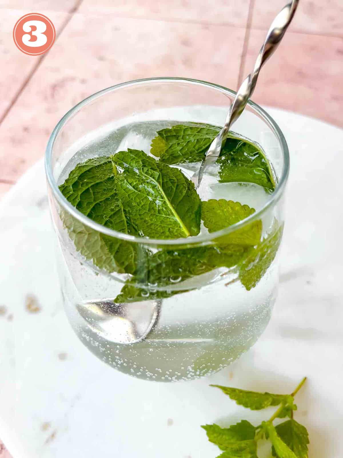 hugo spritz mocktail being muddled with a silver muddler on a marble board labelled number three.