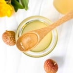 lychee syrup in a glass jar with a wooden spoonful held above it next to fresh lychee and a yellow flower.