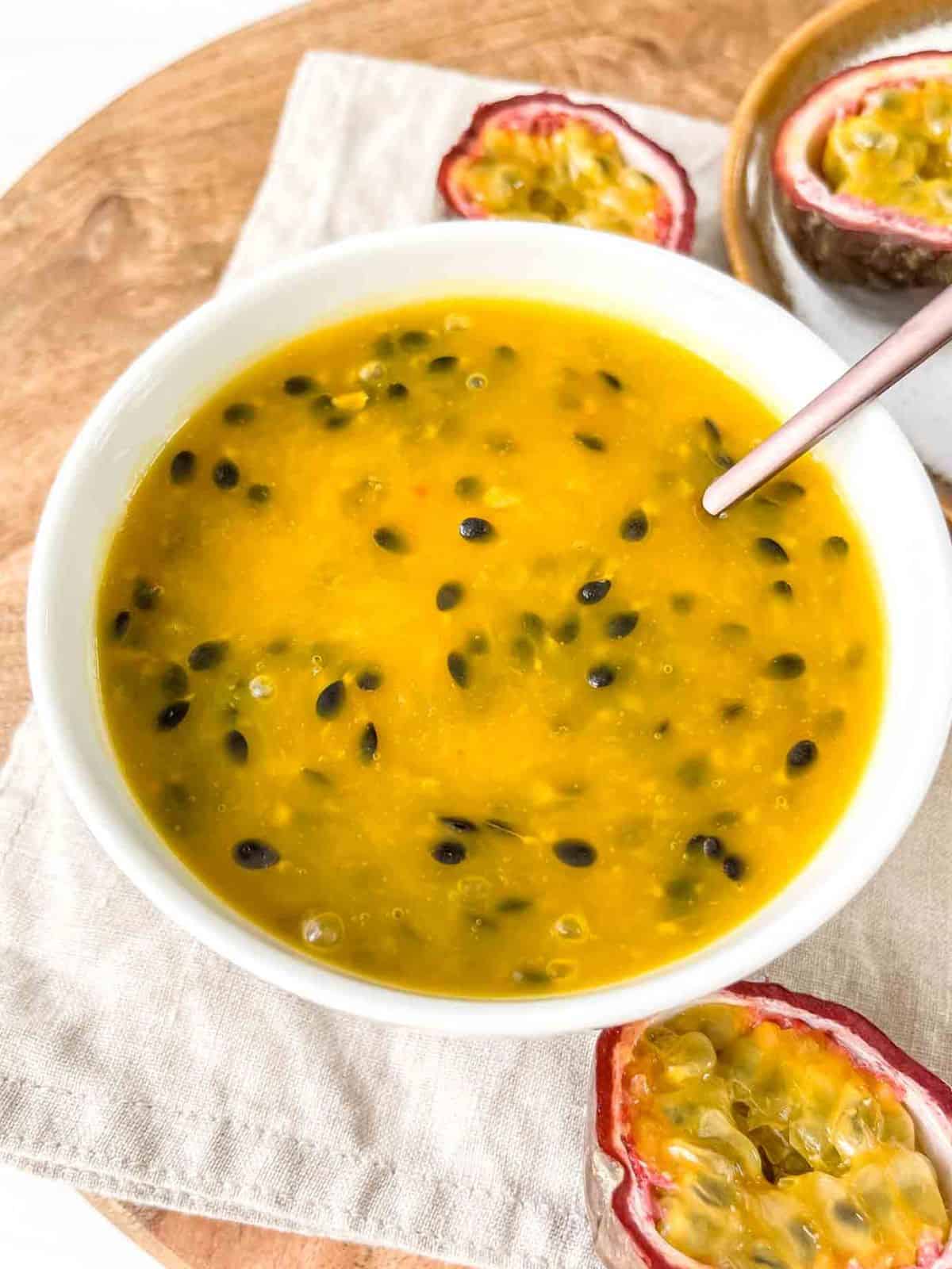 passion fruit coulis in a white bowl with a spoon in it on a wooden board with passion fruit.