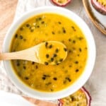 passion fruit coulis in a white bowl with a spoonful held above it on a beige cloth next to passion fruit.