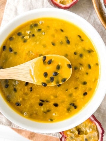 passion fruit coulis in a white bowl with a spoonful held above it on a beige cloth next to passion fruit.