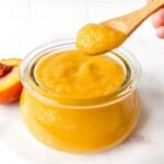 peach applesauce in a glass jar with a spoonful held above it next to half a peach.
