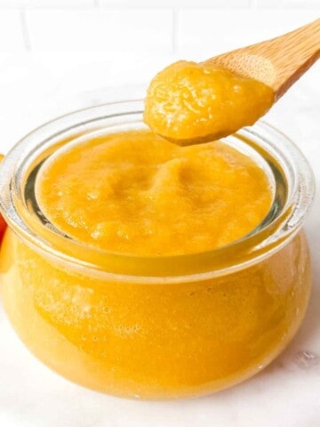 peach applesauce in a glass jar with a spoonful held above it next to half a peach.