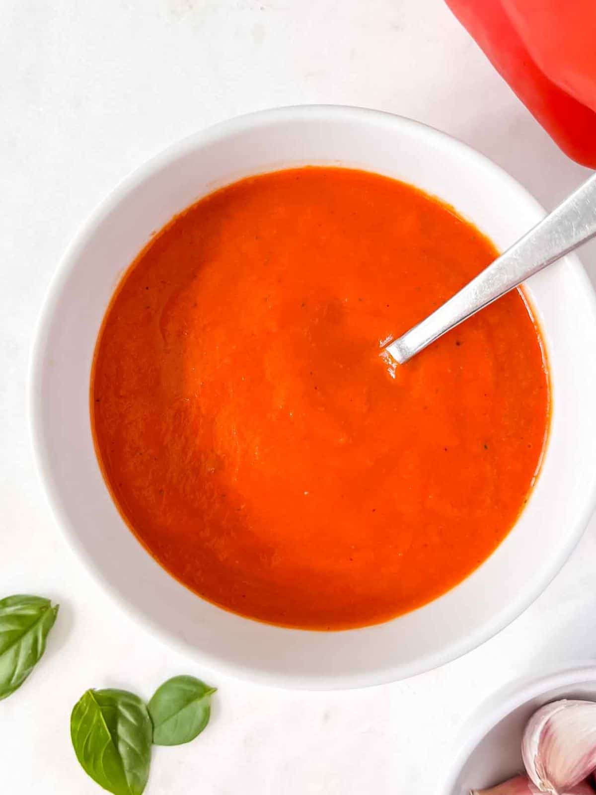 red pepper coulis in a white bowl with a spoon in it next to basil leaves and garlic cloves.