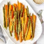 roasted asparagus and carrots on a light grey plate next to a spoon and bowl of sesame seeds.