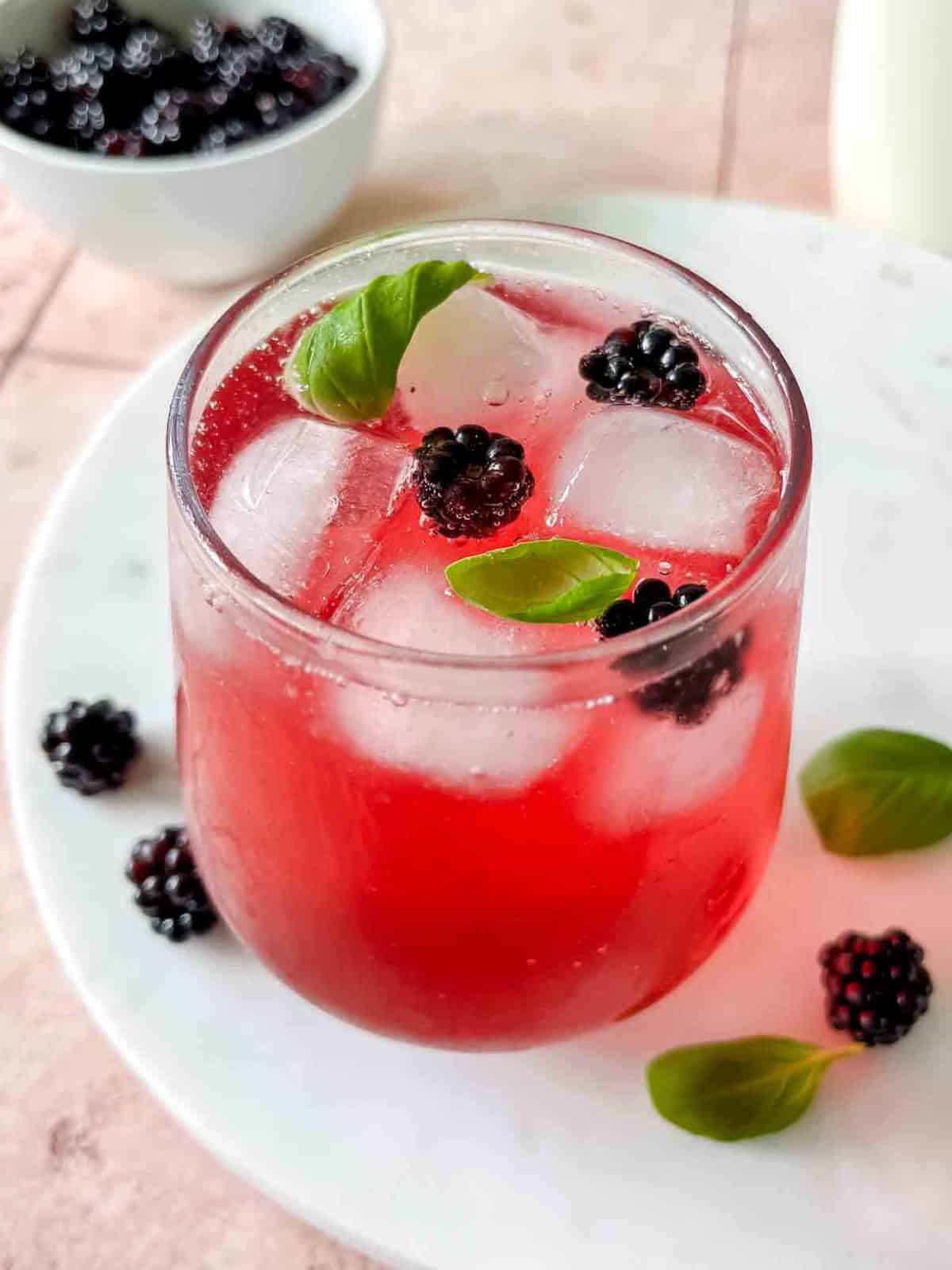 blackberry mocktail in a glass on a white marble board on a pink table next to a bowl of blackberries.