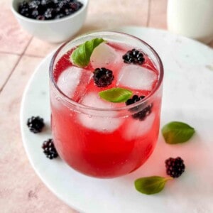 blackberry mocktail in a glass on a white marble board next to a bowl of blackberries and basil leaves.