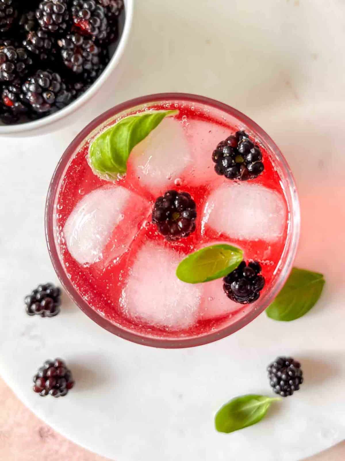blackberry mocktail in a glass next to a bowl of fresh blackberries and basil leaves.