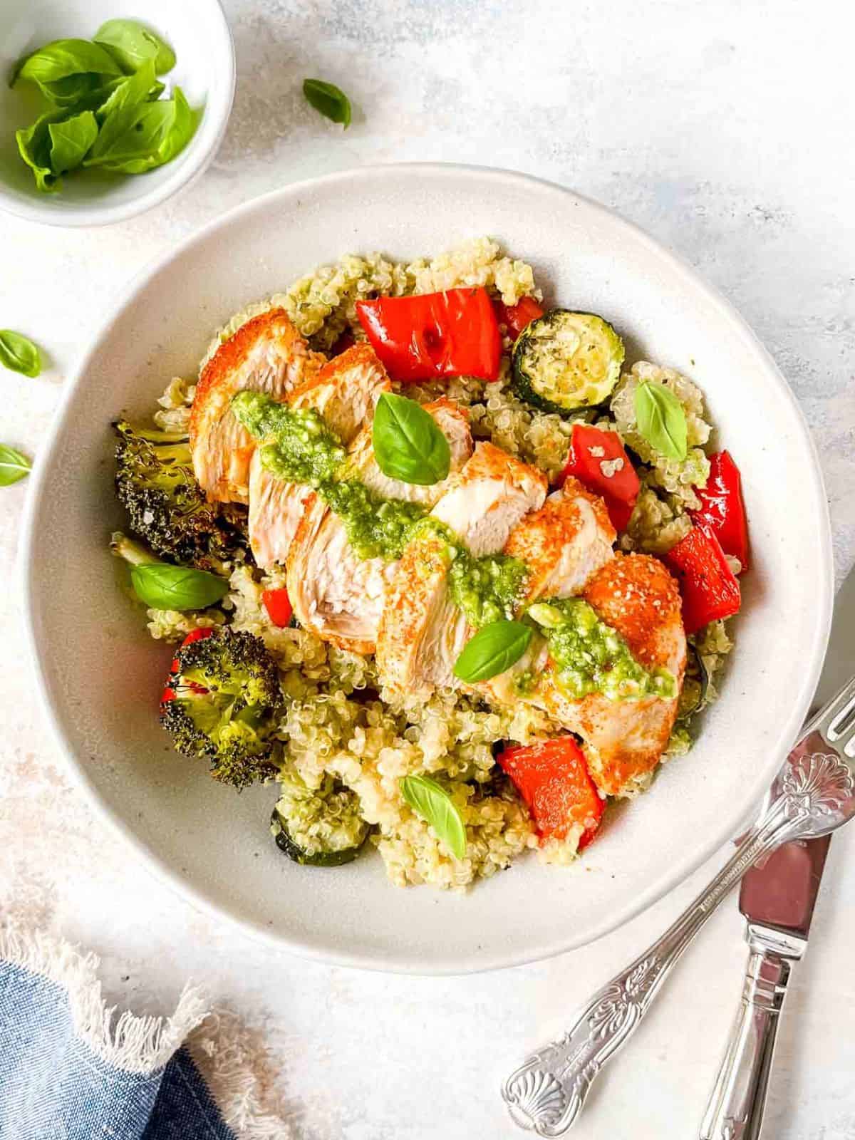 pesto chicken quinoa bowl in a light grey bowl next to cutlery, a blue cloth and small bowl of fresh basil leaves.