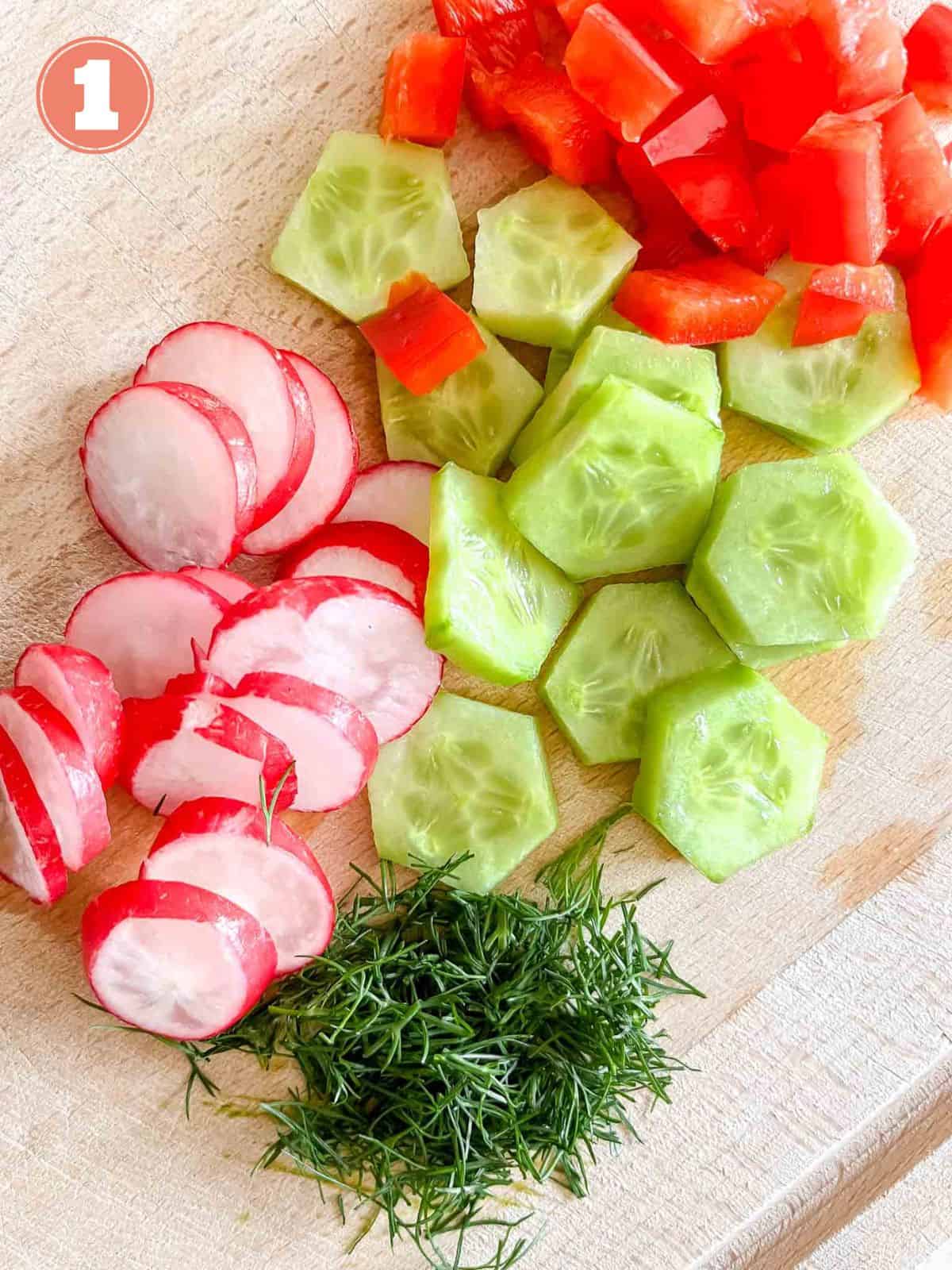 diced radish, dill, cucumber and red bell pepper on a wooden chopping board labelled number one.