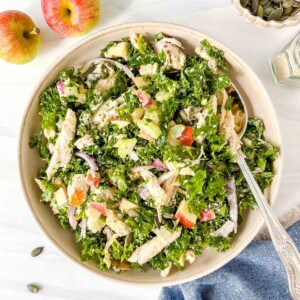 chicken kale salad in a cream bowl with a spoon in it next to red apples and a blue cloth.