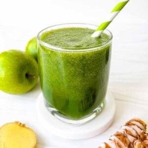 apple ginger smoothie in a glass next to green apples, turmeric and root ginger slices.
