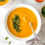 carrot swede soup in a white bowl with a spoon in it next to small bowls of carrots and parsley.