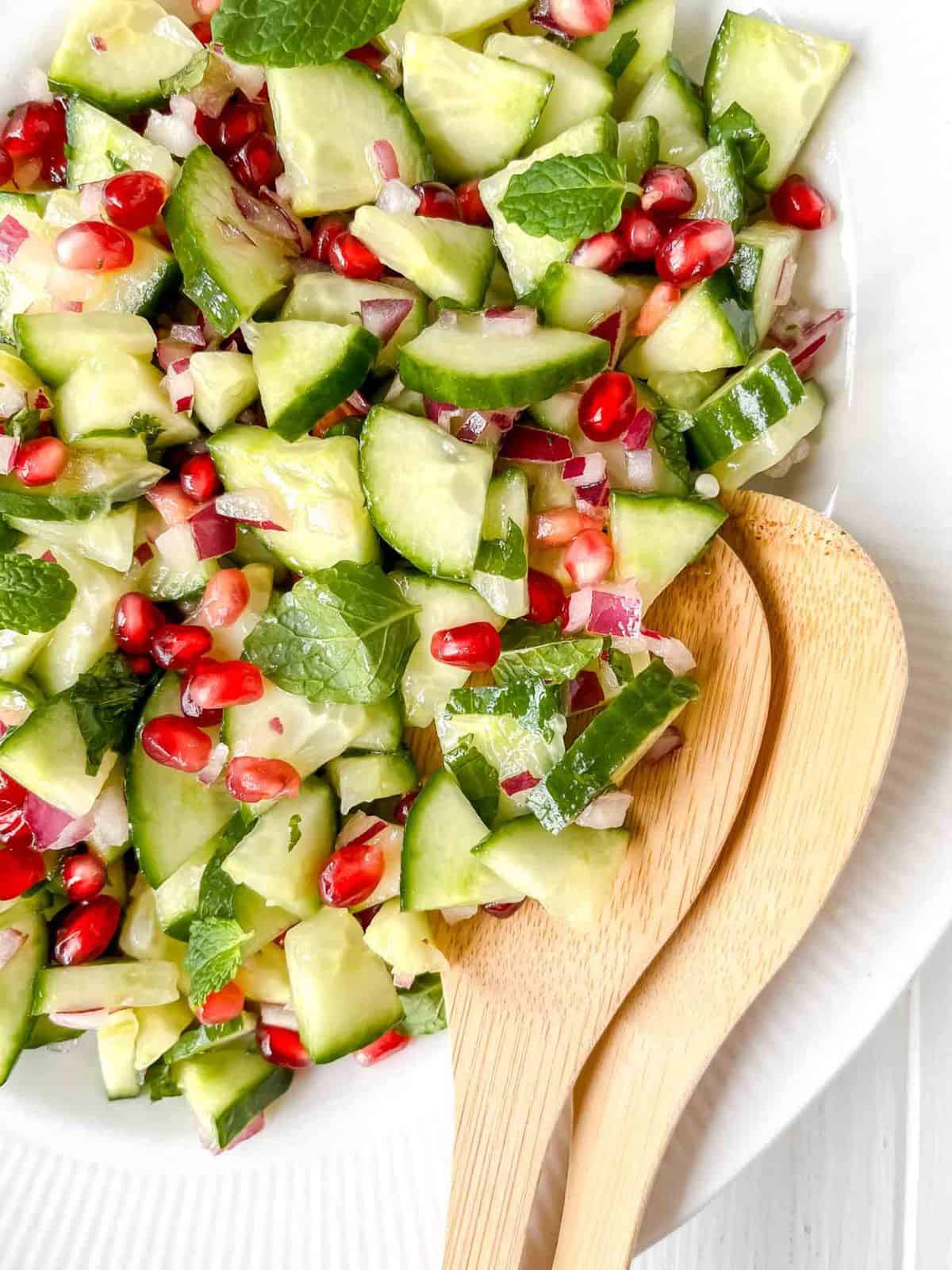 cucumber pomegranate salad on a white plate with wooden salad servers.