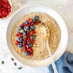 quinoa flake porridge in a cream bowl topped with pomegranate seeds and blueberries.