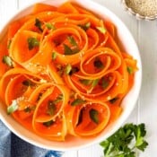 raw carrot salad in a white bowl sprinkled with fresh parsley and sesame seeds on a blur cloth.