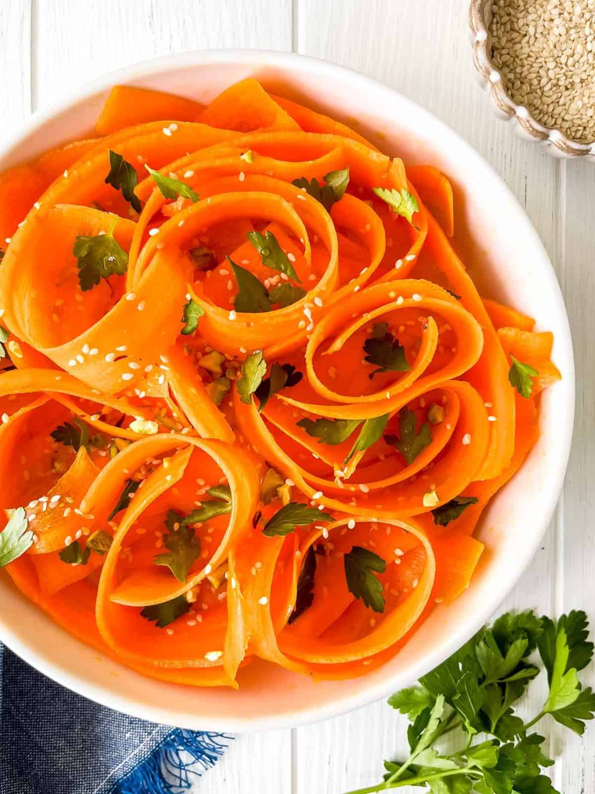 raw carrot salad in a white bowl sprinkled with sesame seeds and parsley next to a bowl of sesame seeds.