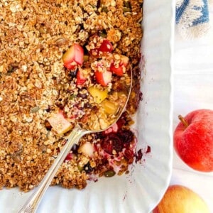 apple blueberry crisp in a baking dish with a spoon next to red apples.
