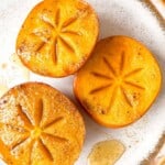 three halved baked persimmons on a cream plate drizzled with honey and spices.