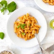 red pepper pesto pasta on two white plates next to basil leaves, olive oil and pine nuts.