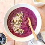 smoothie bowl without banana in a white bowl topped with granola, coconut and seeds on a pink background.
