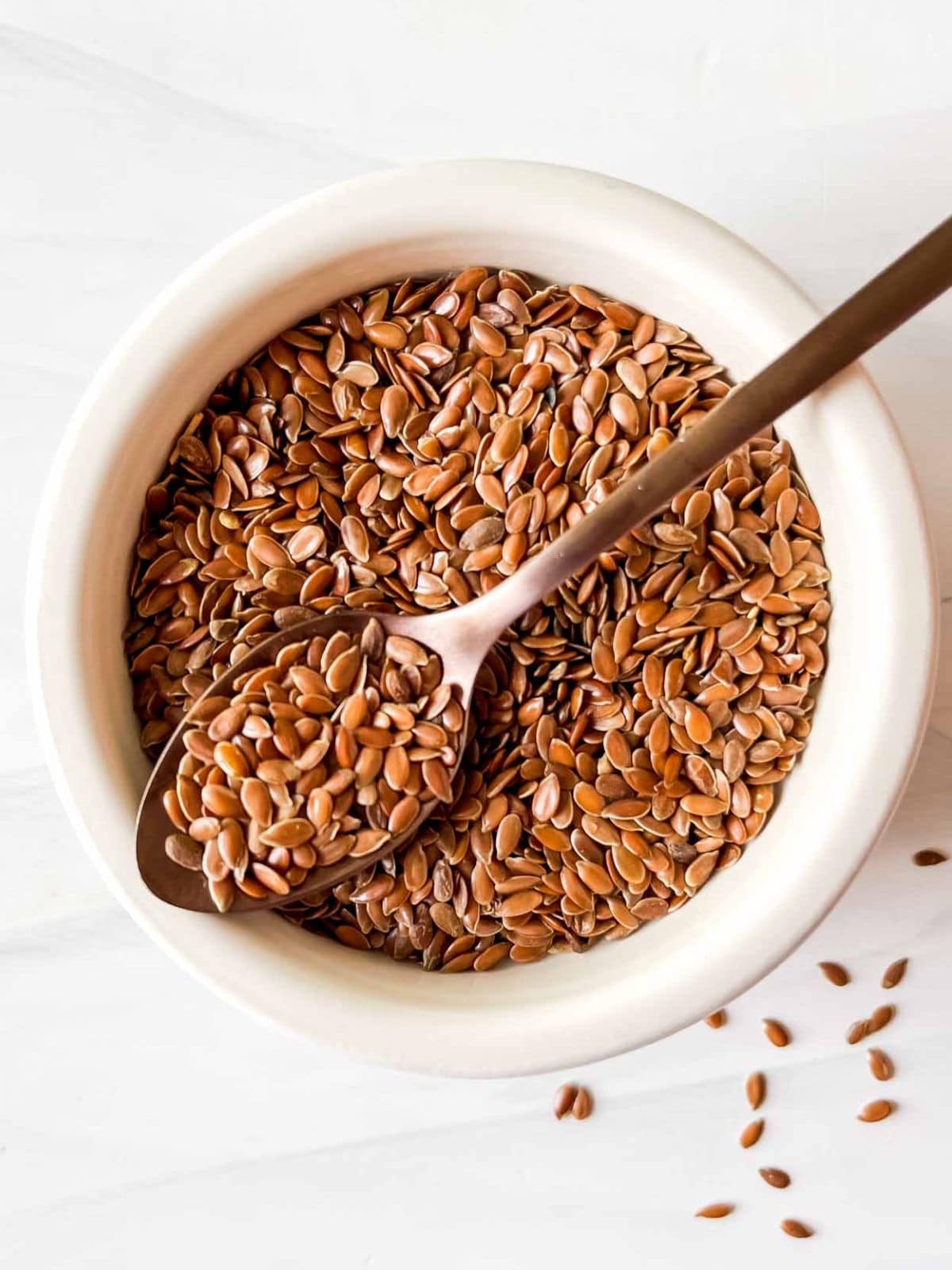 flaxseeds in a white bowl with a metal spoonful of seeds on top.