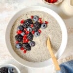 hemp chia pudding topped with berries in a white bowl with a spoon next to a blue cloth.