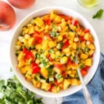 peach corn salsa in a white bowl with a spoon next to fresh peaches and cilantro.