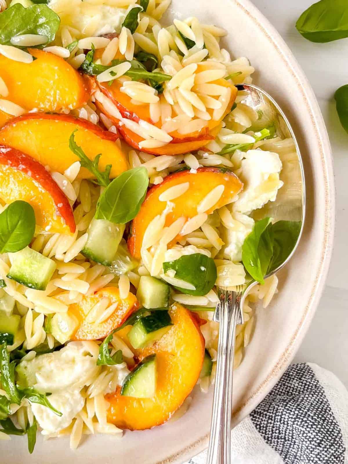 peach pasta salad in a cream bowl with a spoon next to basil leaves.