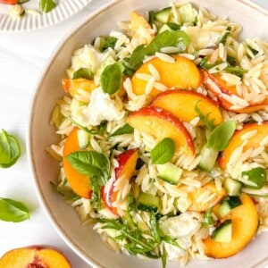 peach pasta salad in a cream bowl next to basil leaves and a fresh peach.