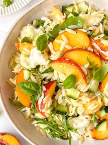 peach pasta salad in a cream bowl next to basil leaves and a fresh peach.