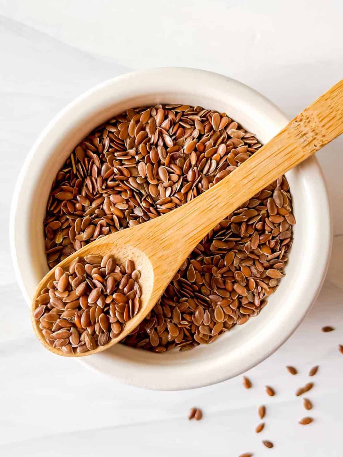 flax seeds in a white bowl with a wooden spoonful of seeds on top.
