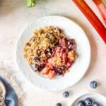 blueberry rhubarb crisp on a white plate next to rhubarb stems and a bowl of blueberries.