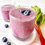 rhubarb smoothie in two glasses next to rhubarb stems and blueberries.