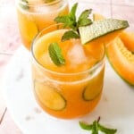 cucumber melon agua fresca in two glasses topped with mint leaves, next to slices of cantaloupe melon.
