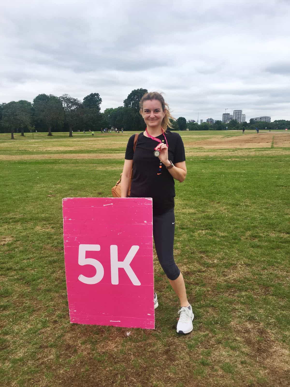 woman holding a medal in a park next to a 5 km sign.