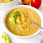 celery apple soup in a white bowl next to red apples and celery leaves.