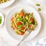 vegetable pesto pasta on a white plate with a fork next to fresh basil leaves.