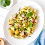 cucumber potato salad with dill and radishes on a white plate next to a small bowl of fresh mint leaves and blue cloth.